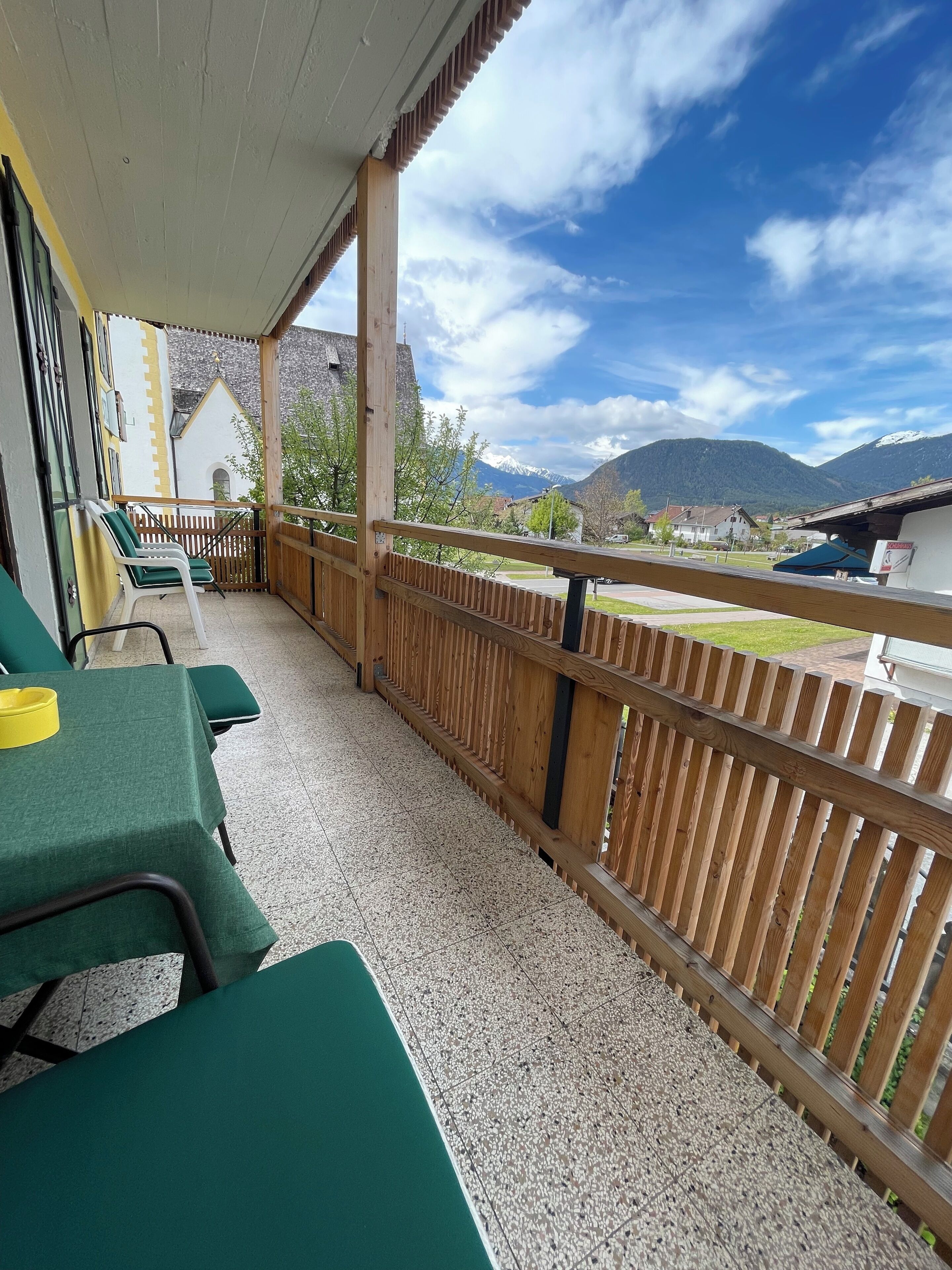 Un'area salotto esterna coperta con sedie e tavoli verdi, ringhiere in legno e vista sulle montagne e sugli edifici sullo sfondo.