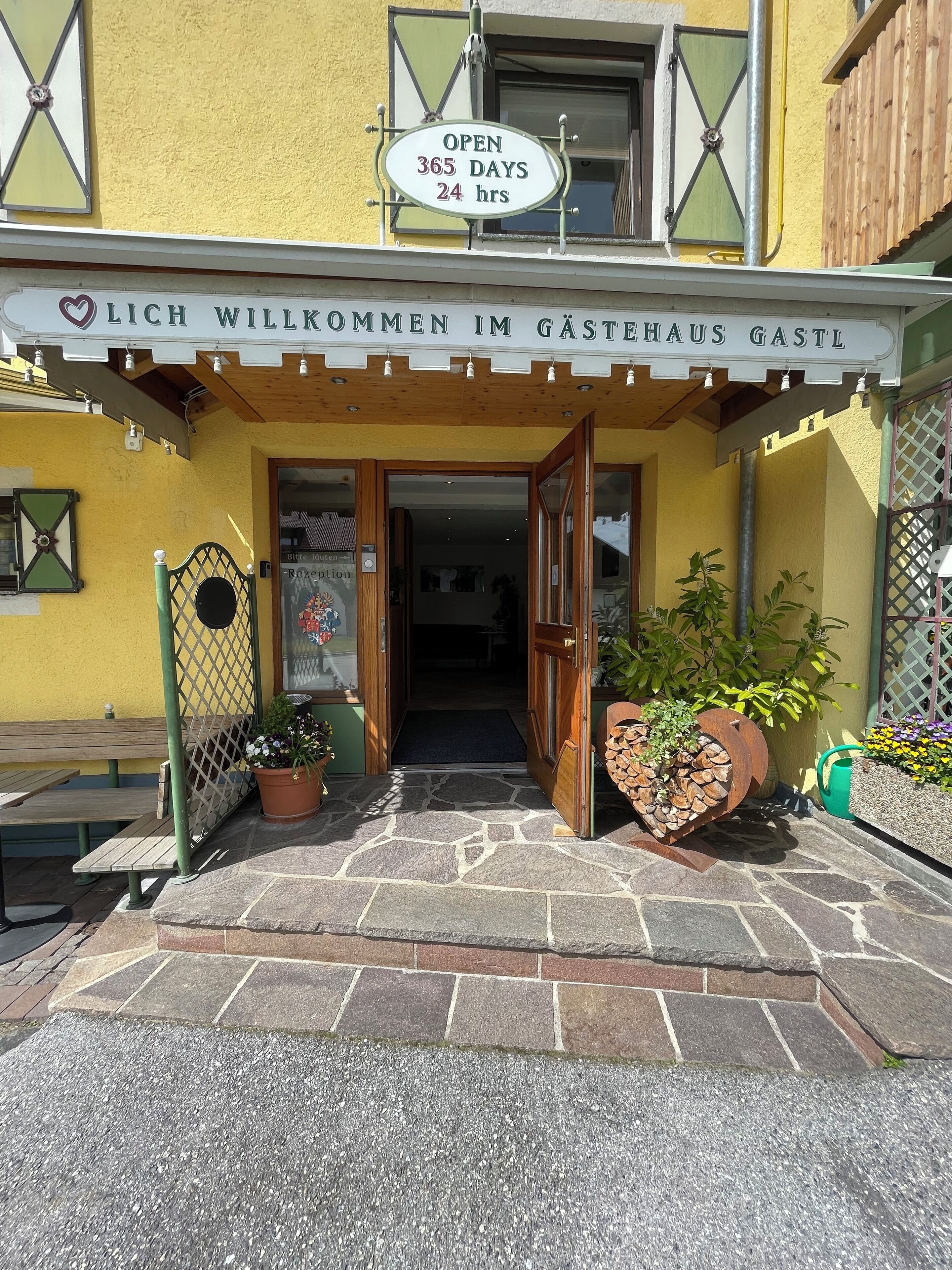 Ein Gebäudeeingang mit einem Schild, auf dem steht: 'GEÖFFNET 365 TAGE 24 Stunden' und 'HERZLICH WILLKOMMEN IM GÄSTERHAUS GASTL'.
