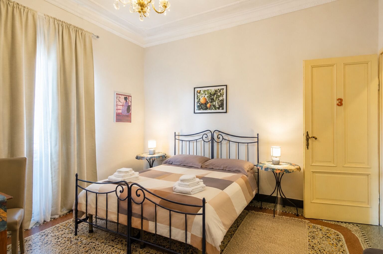A bedroom with a bed, two bedside tables, a chair, a door, and a chandelier.