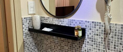 Un baño con lavamanos blanco, un espejo, un secador de pelo y un estante con artículos de aseo.