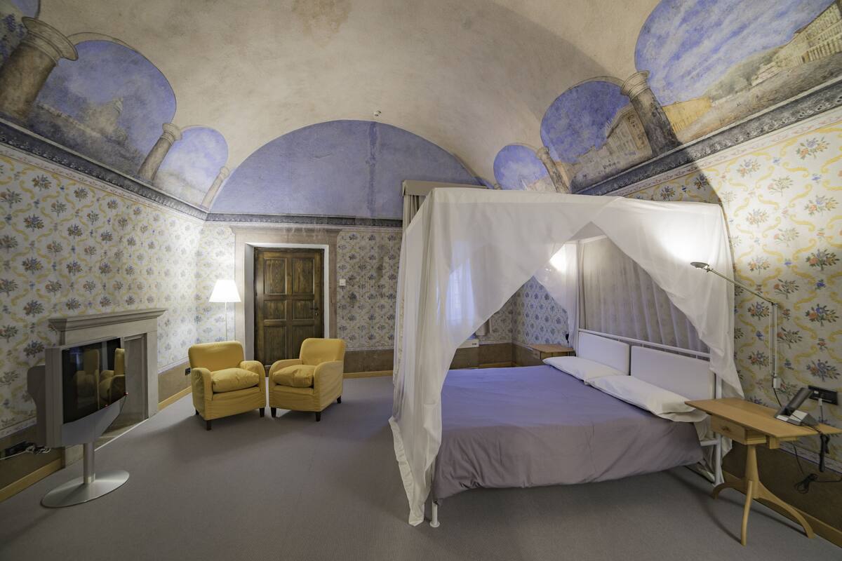 A room with a four-poster bed, a television, two yellow armchairs, and a fireplace.