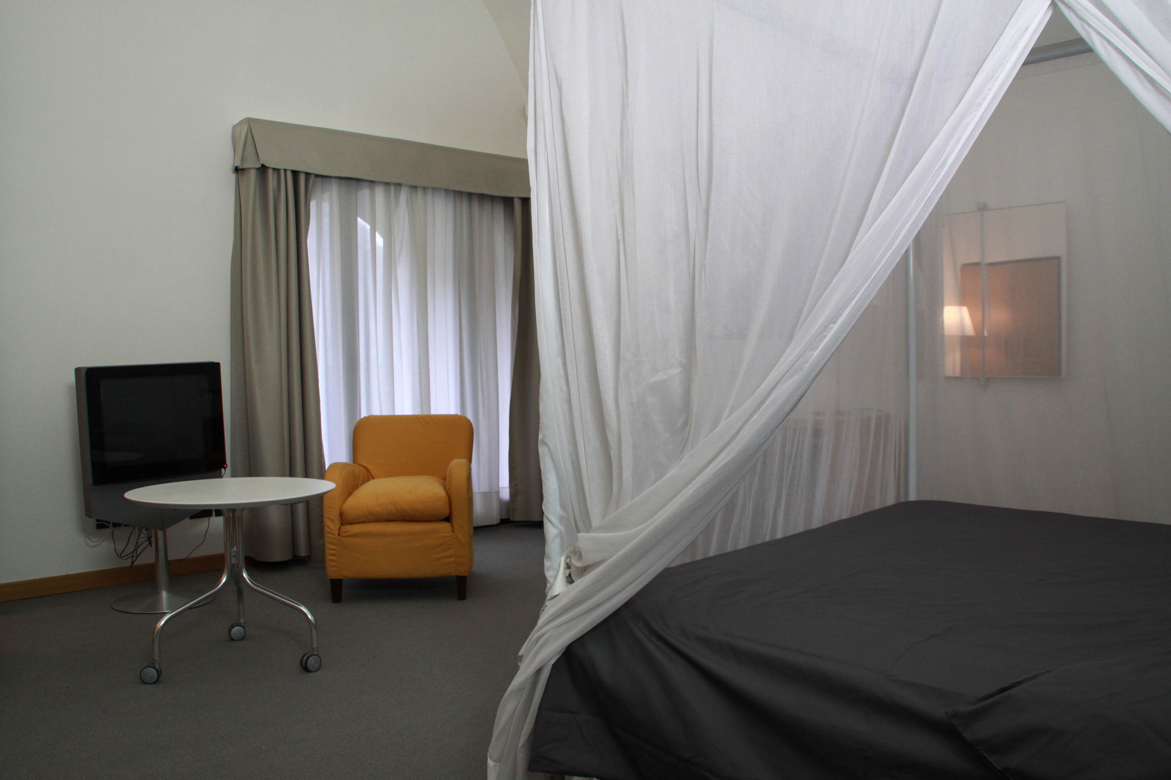 A hotel room with a bed, a television, a small table, an armchair, and a window with curtains.