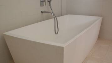 A modern, rectangular bathtub with a handheld showerhead, set against a plain tiled wall.