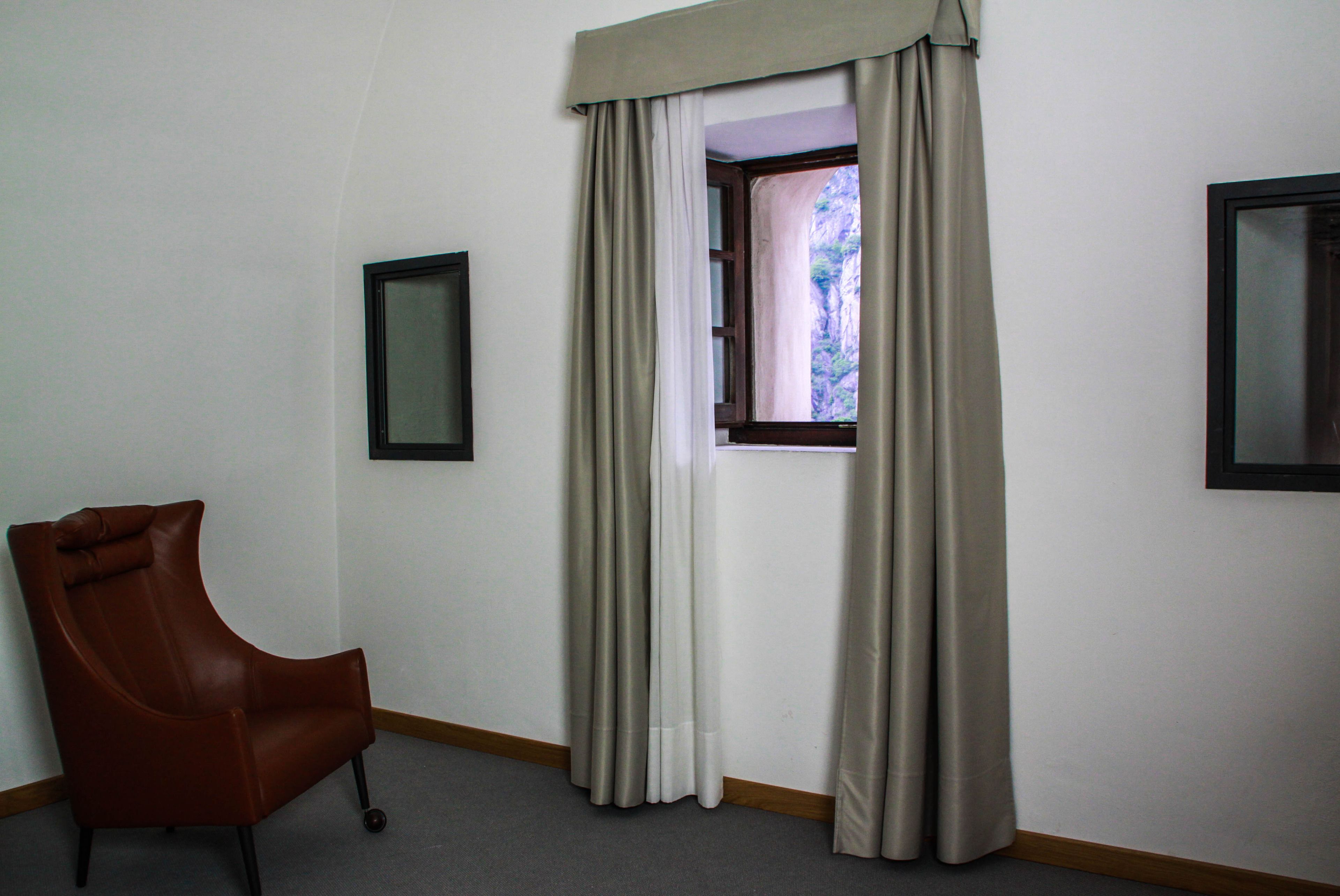 A room with a single brown leather chair, a window with beige curtains, and two framed pictures on the walls.