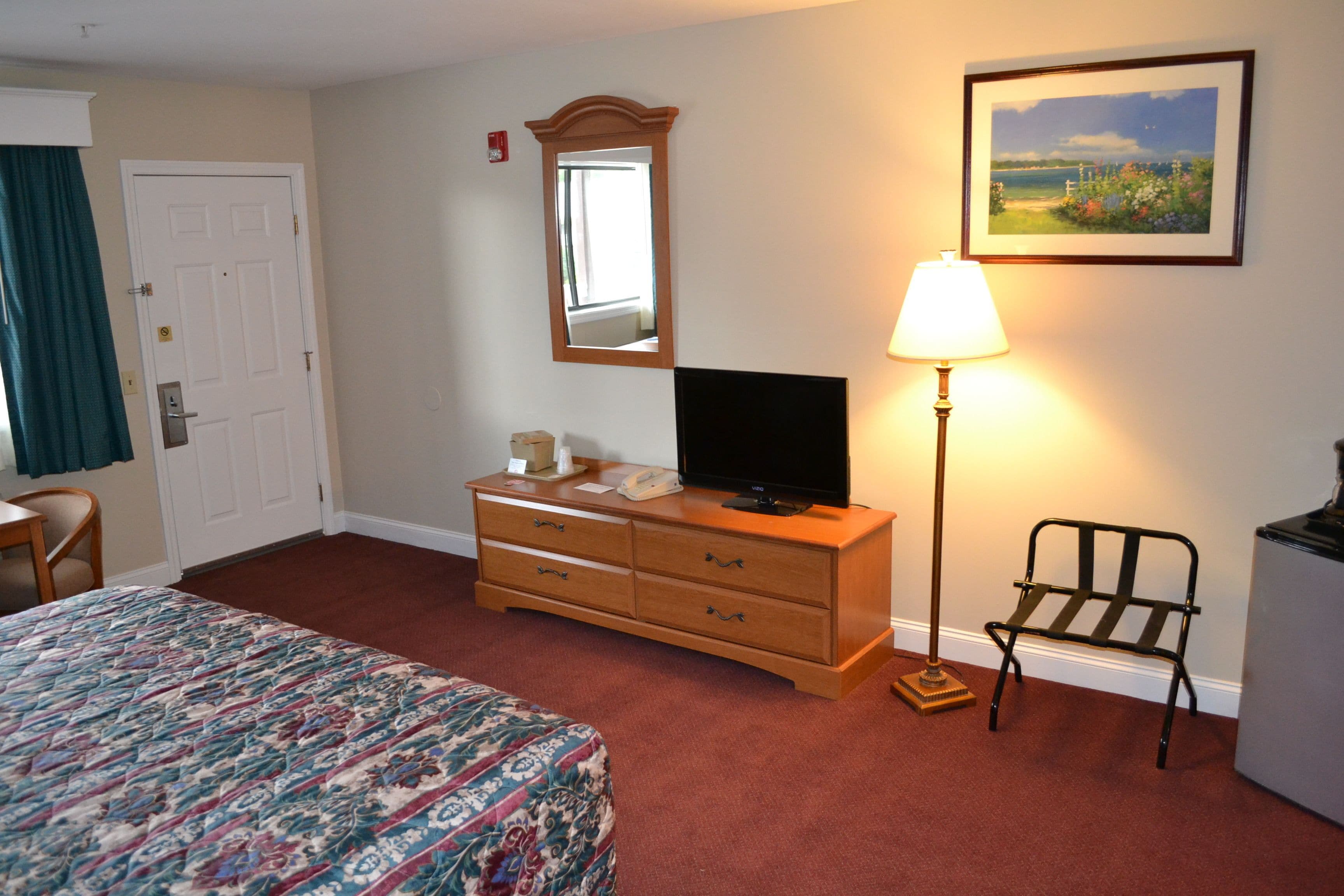 A hotel room with a bed, a dresser with a television, a lamp, a chair, and a painting on the wall.
