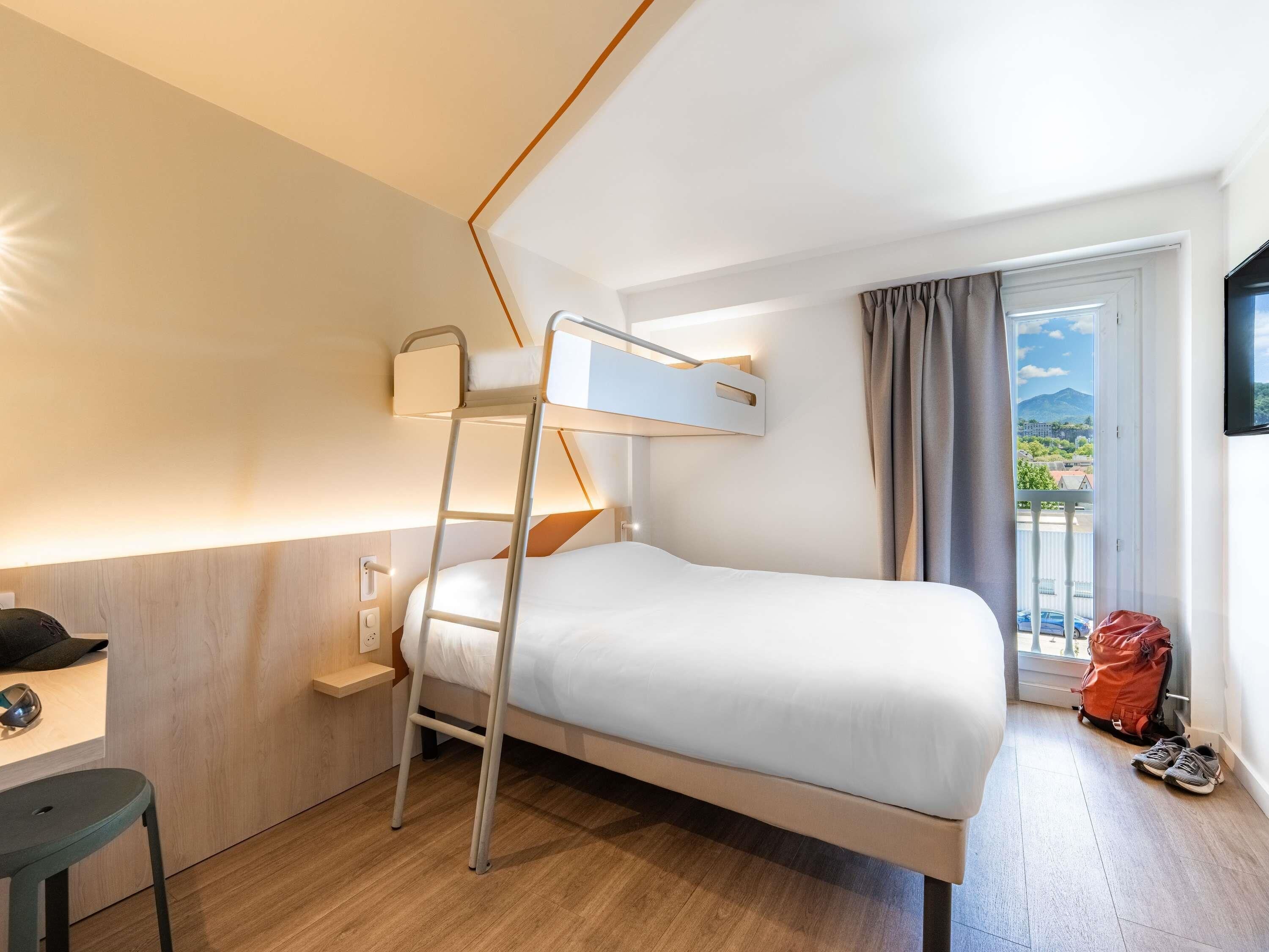 A compact hotel room with a bunk bed, a desk with a chair, a window with curtains, and a view of the outside.