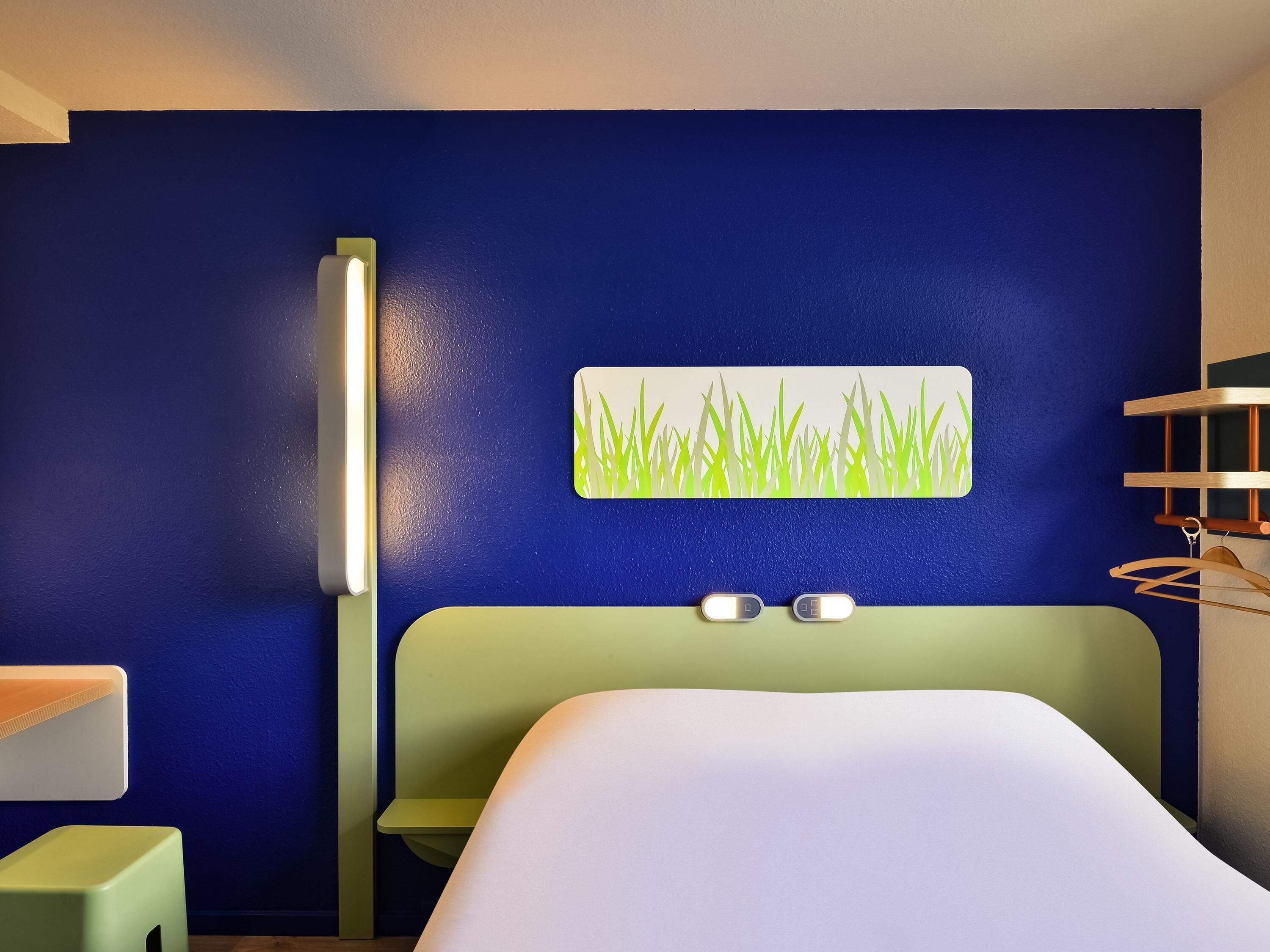 A hotel room with a bed, a wall-mounted light, a small table, and a wall art piece.