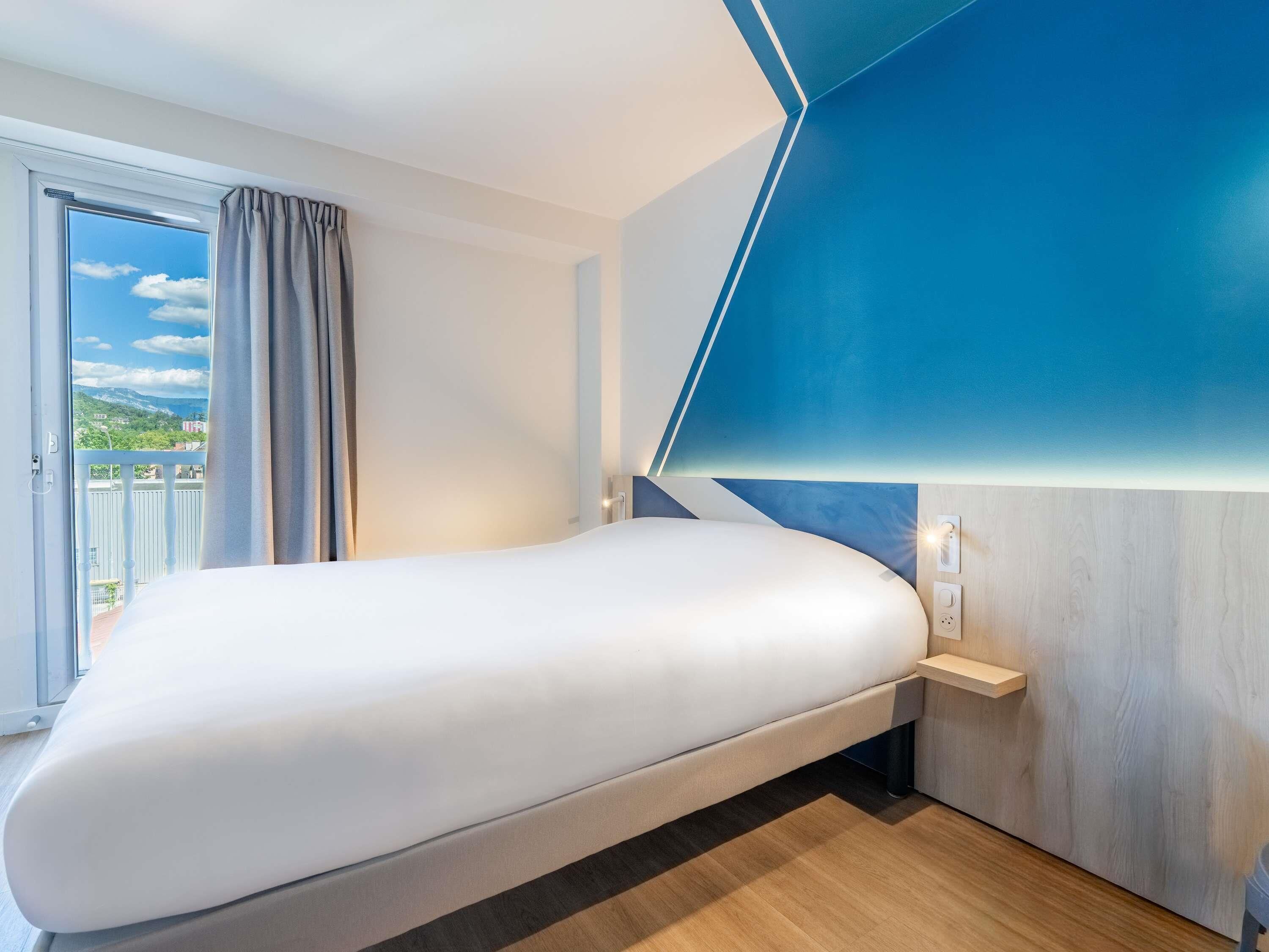 Une chambre d’hôtel moderne dotée d’un grand lit, offrant une vue sur l’extérieur et agrémentée d’un mur d’accent d’une couleur bleue originale.