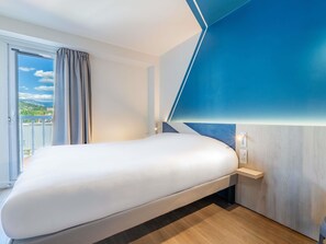 A modern hotel room with a large bed, a view of the outdoors, and a unique blue accent wall. - ibis budget Chambéry Centre Ville - Hotel Rénové (Chambery)