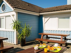 A seaside patio with wooden benches and tables, a potted plant, and a breakfast spread including croissants, fruit, and juice. - ibis budget Chambéry Centre Ville - Hotel Rénové (Chambery)
