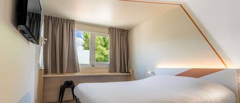 Un dormitorio con cama, televisor montado en la pared, ventana con cortinas y lámpara.