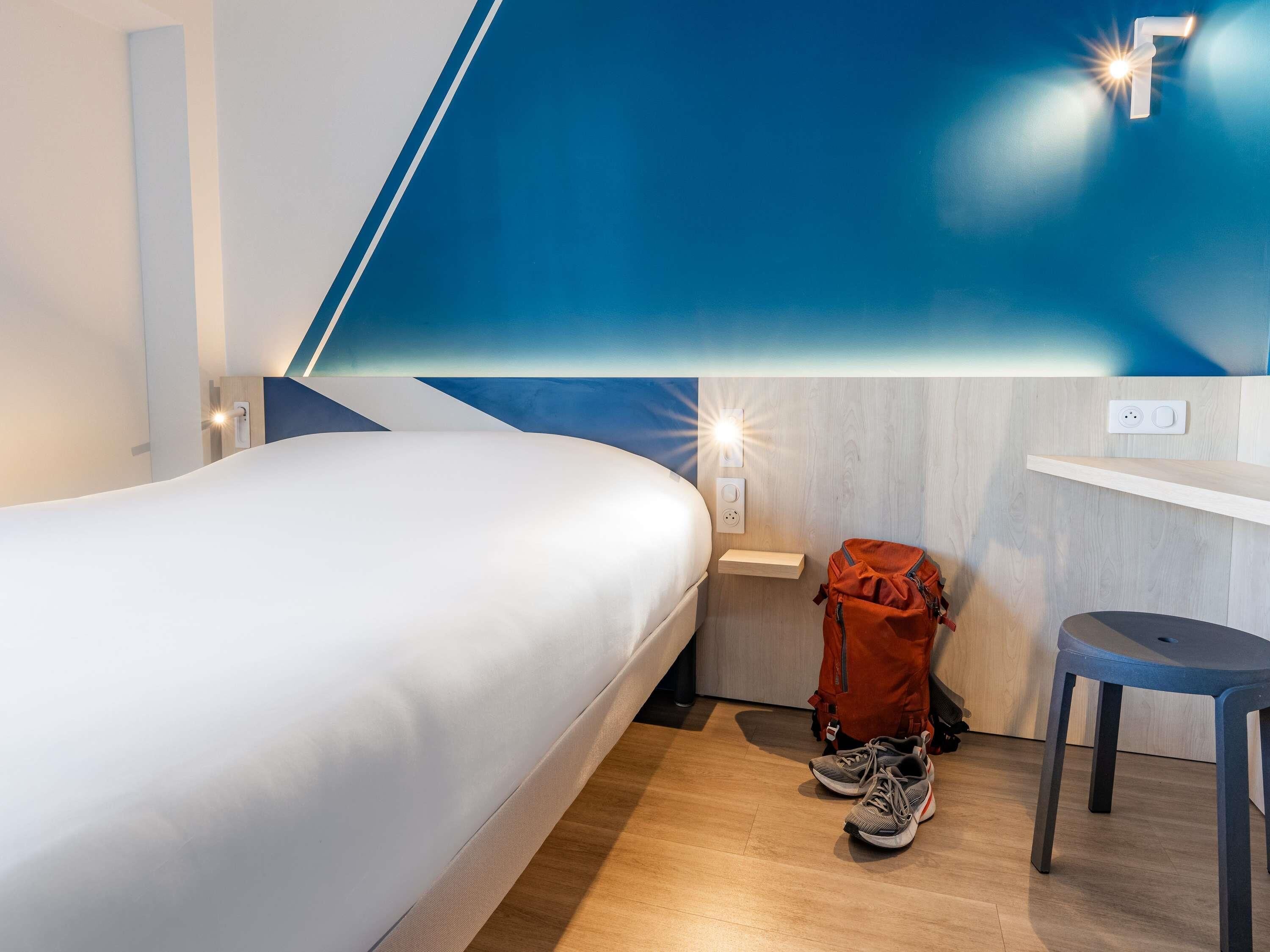 Una habitación de hotel con cama, mesita de noche, lámpara en la pared, suelo de madera y una mochila roja.