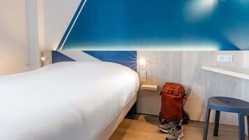 Una habitación de hotel con cama, mesita de noche, lámpara en la pared, suelo de madera y una mochila roja.