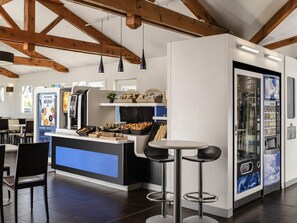 A modern café with a central counter, bar stools, a coffee machine, and a refrigerated display. - ibis budget Chambéry Centre Ville - Hotel Rénové (Chambery)