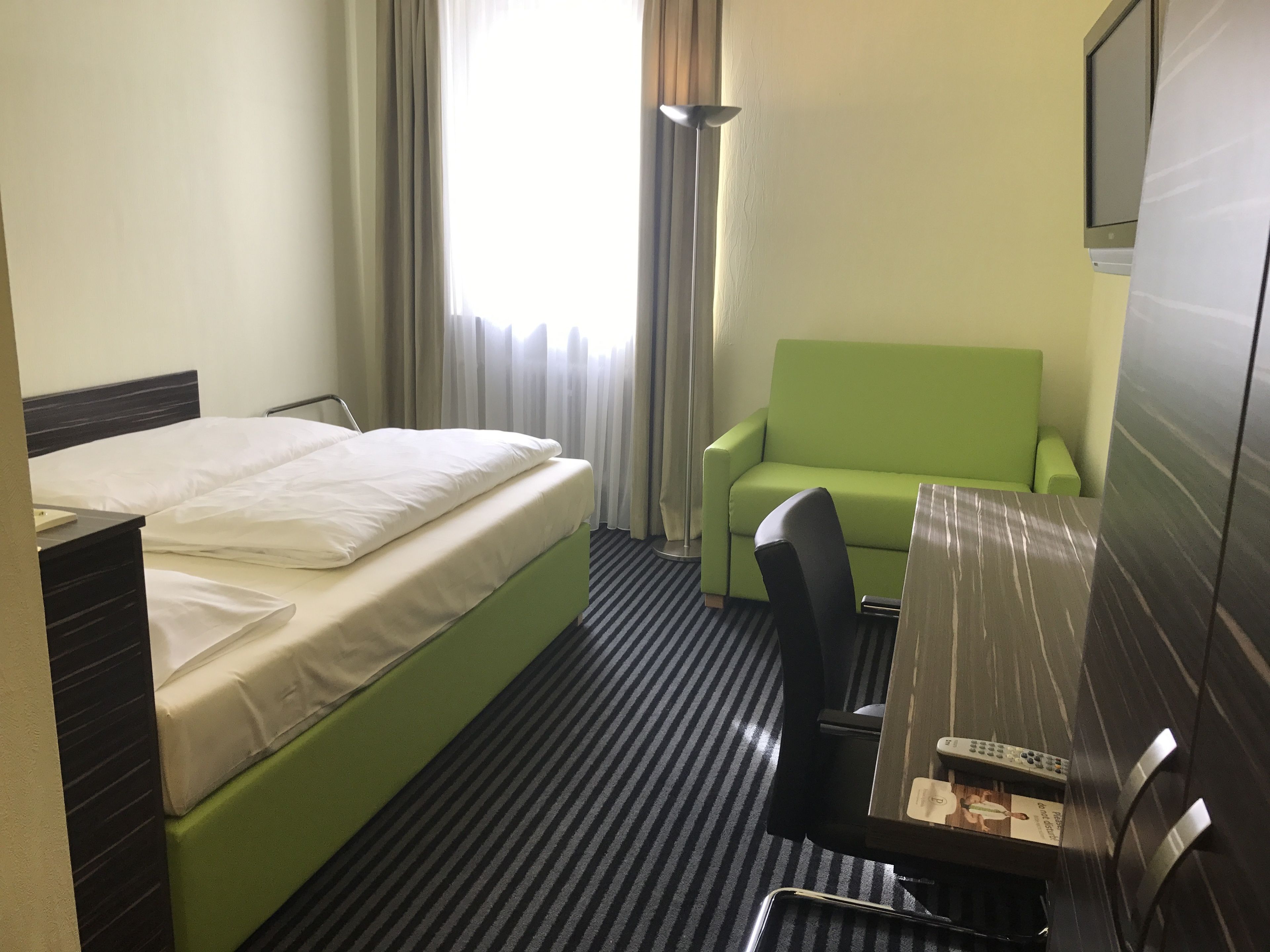 A hotel room with a bed, a green sofa, a desk with a chair, a TV, and a lamp.