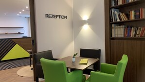 A reception area with a 'RECEPTION' sign, a wooden table, green chairs, a bookshelf, and a small plant on the table.