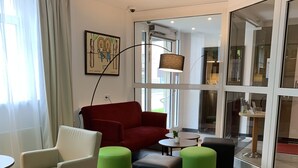 A modern hotel room with a red sofa, a white armchair, and green ottomans. There is a small round table with a plant, and a large window with curtains.
