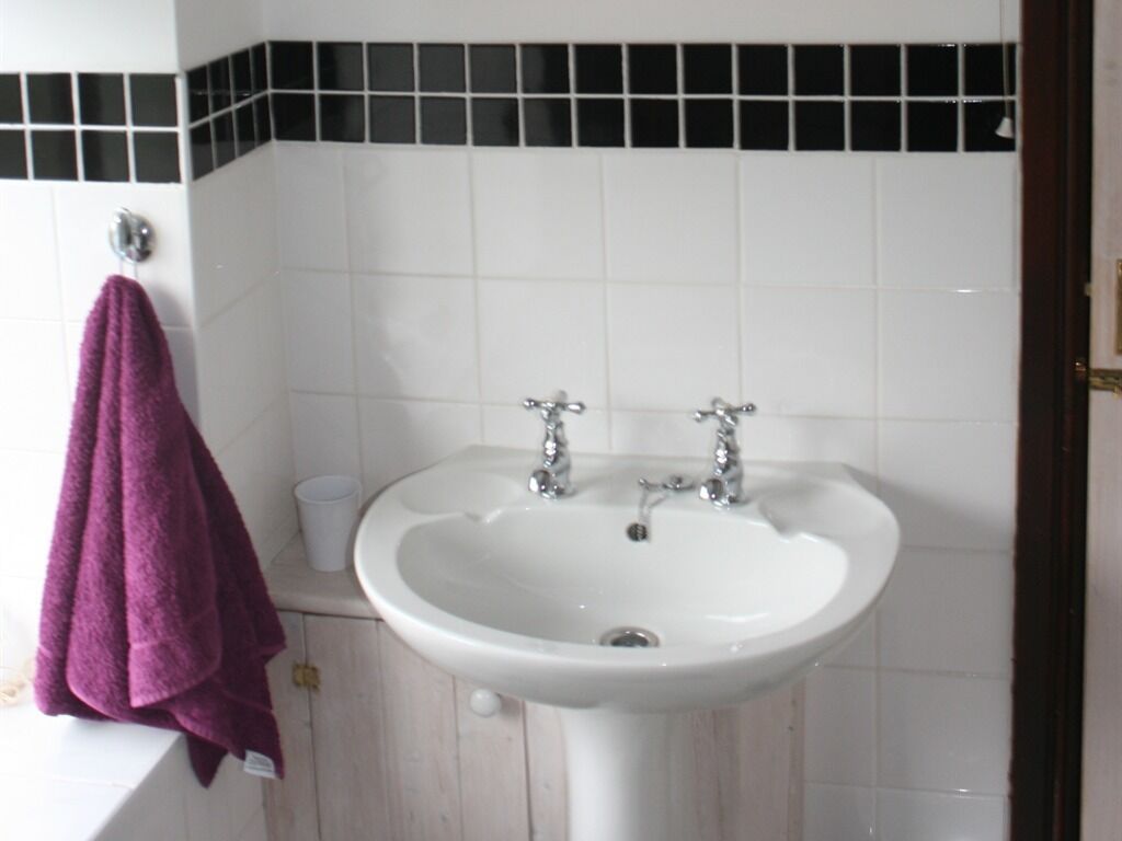 Ein Badezimmer mit einem weißen Waschbecken, zwei Wasserhähnen, einem Spiegel und einem violetten Handtuch, das an der Wand hängt.