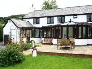 A white house with a large porch, a conservatory, and a garden with flowers and a bench. - The Old School House (Llanbrynmair)