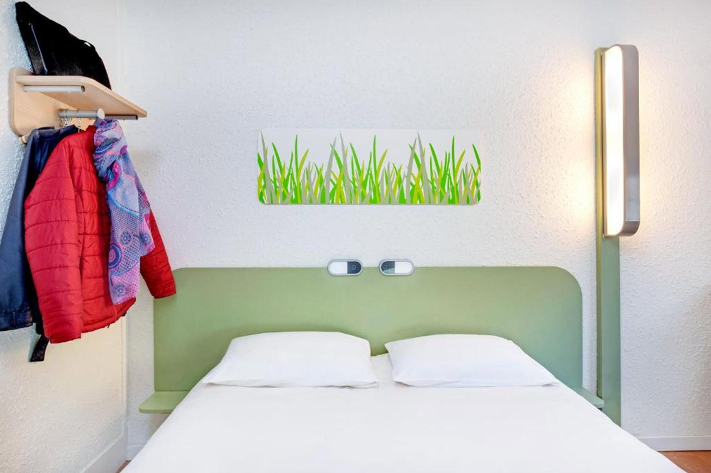Quarto de hotel com cama verde, almofadas brancas, luminária na parede e pintura de grama.