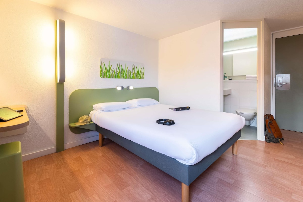 Une chambre d’hôtel avec un lit, une table de chevet, une lampe et une salle de bain visible à travers une porte ouverte.