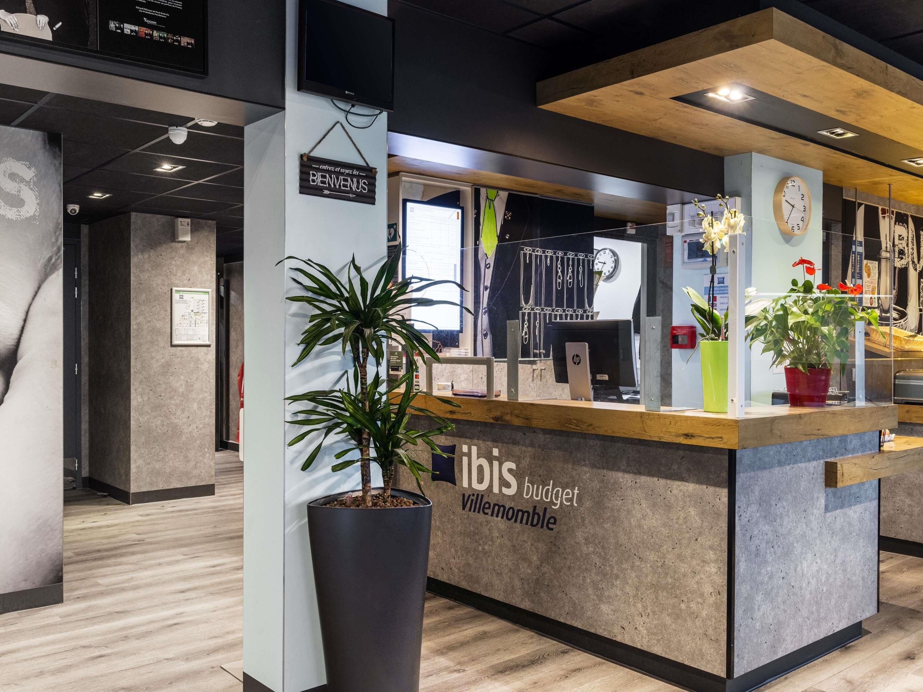 Una recepción con un cartel que dice 'ibis budget Villermontble', una planta en maceta grande, un reloj y un monitor de computadora.