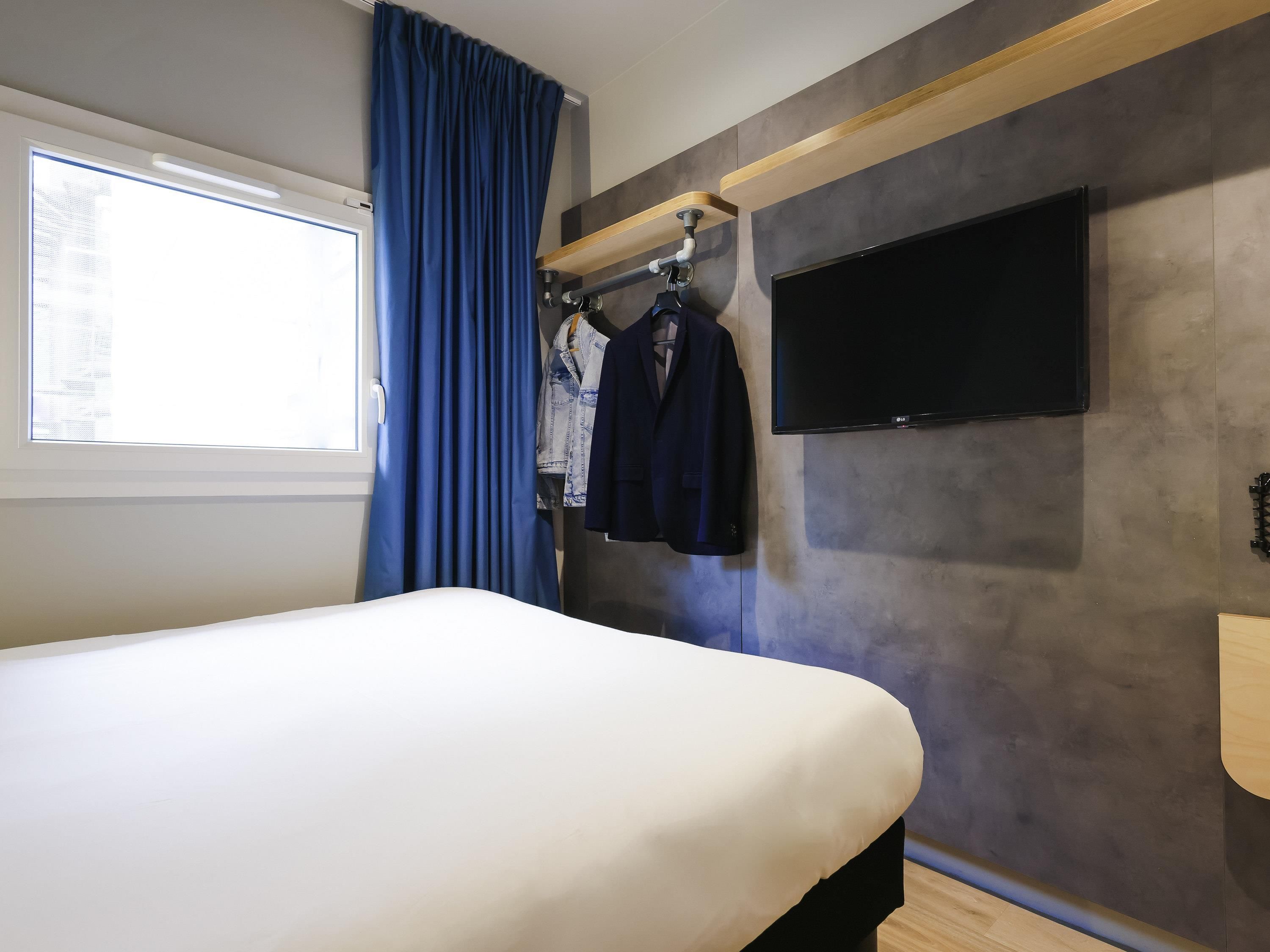 Una habitación de hotel con una cama, un televisor montado en la pared, una ventana con cortinas y un perchero con chaquetas.
