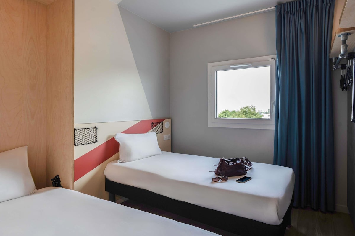 A hotel room with two beds, a window with curtains, and a view of greenery outside.