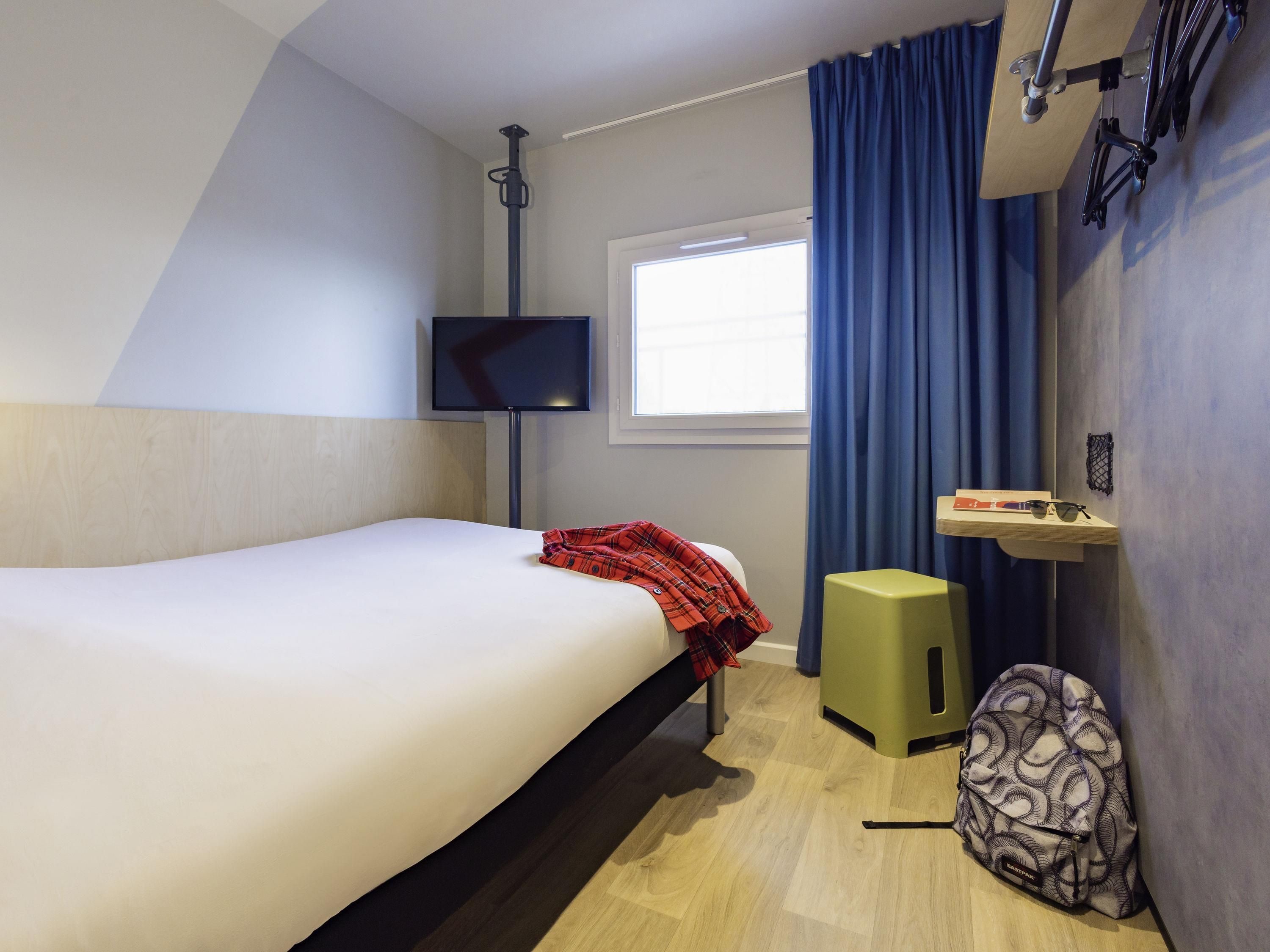 Quarto de hotel com cama, televisão, janela com cortinas, prateleira pequena com objetos e uma mochila.