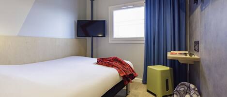A hotel room with a bed, a television, a window with curtains, a small shelf with items, and a backpack.