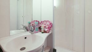 Une salle de bain avec un lavabo blanc, un miroir et des carreaux blancs. On peut voir des articles de toilette sur le lavabo et un espace douche.