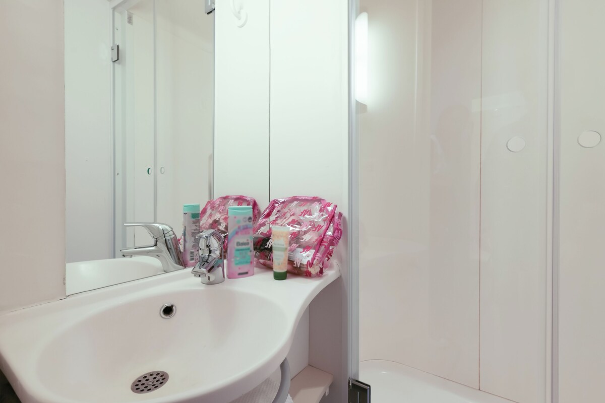 A bathroom with a white sink, mirror, and white tiles. There are toiletries on the sink and a shower area visible.