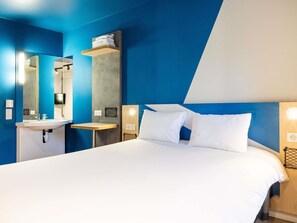 A modern hotel room with a large bed, a bedside table, a built-in shelf with towels, a mirror, and a wall-mounted light. - ibis budget Villemomble (Villemomble)