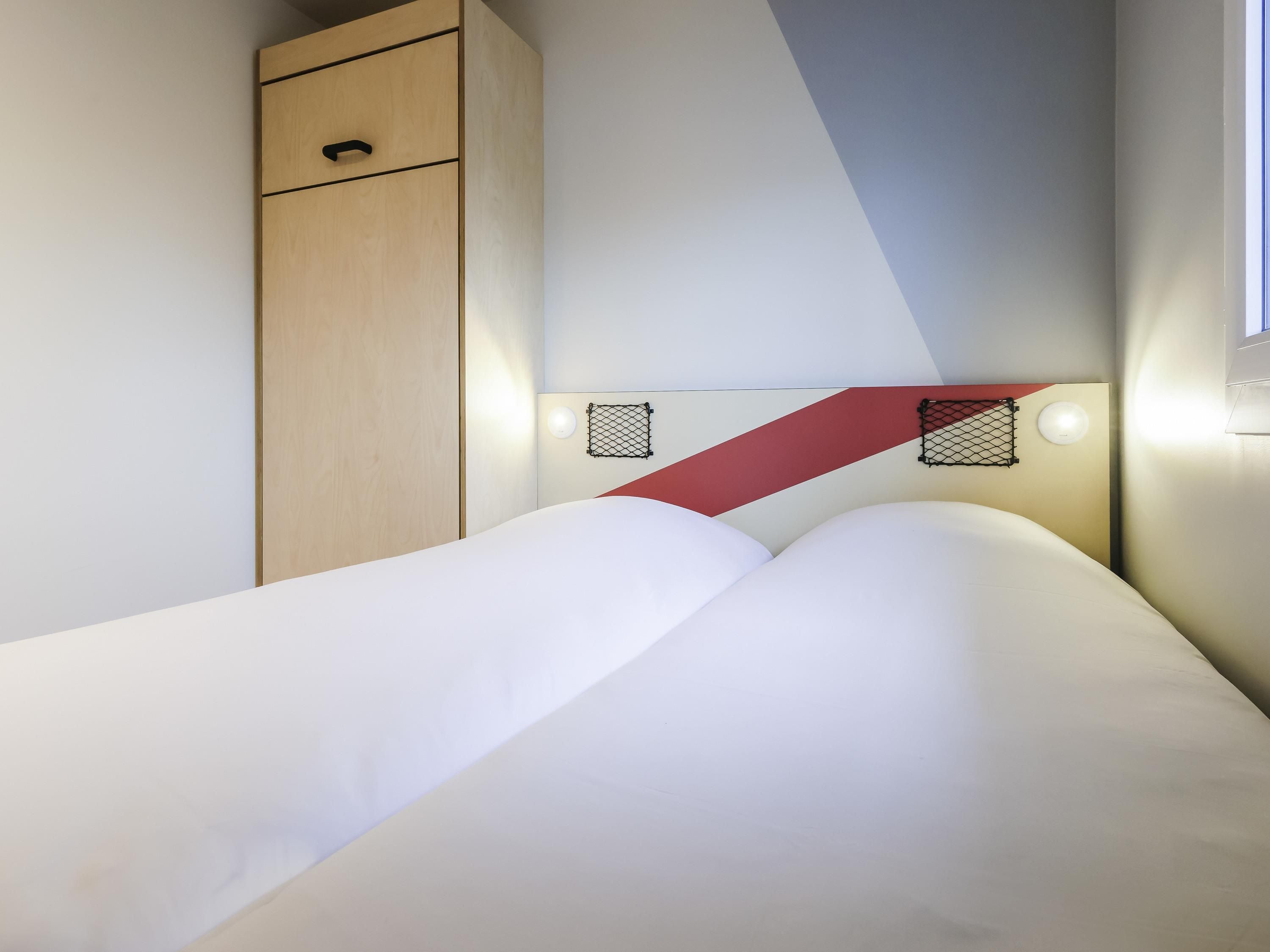 A bed with white bedding, a wooden wardrobe, and a red diagonal accent on the wall.
