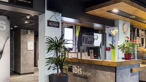 A reception desk with a sign reading 'ibis budget Villermontble,' a large potted plant, a clock, and a computer monitor. - ibis budget Villemomble (Villemomble)