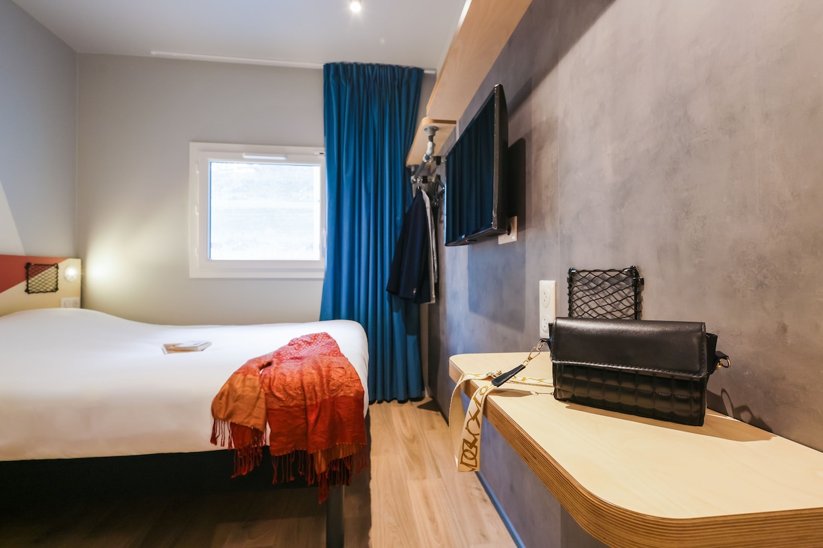 A hotel room with a bed, a desk, a TV, and a window with curtains.