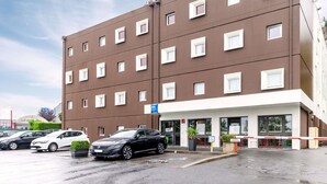 Un hôtel à plusieurs étages, doté d’une façade marron, de nombreuses fenêtres et d’un parking où sont stationnées plusieurs voitures.