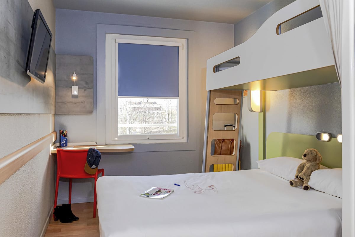 Een compacte hotelkamer met een stapelbed, een bureau met stoel, een raam met uitzicht en een teddybeer op het bed.