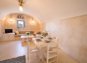 A dining area with a table set for four, a sofa with cushions, a television on a cabinet, and a view of the outdoors through a window.