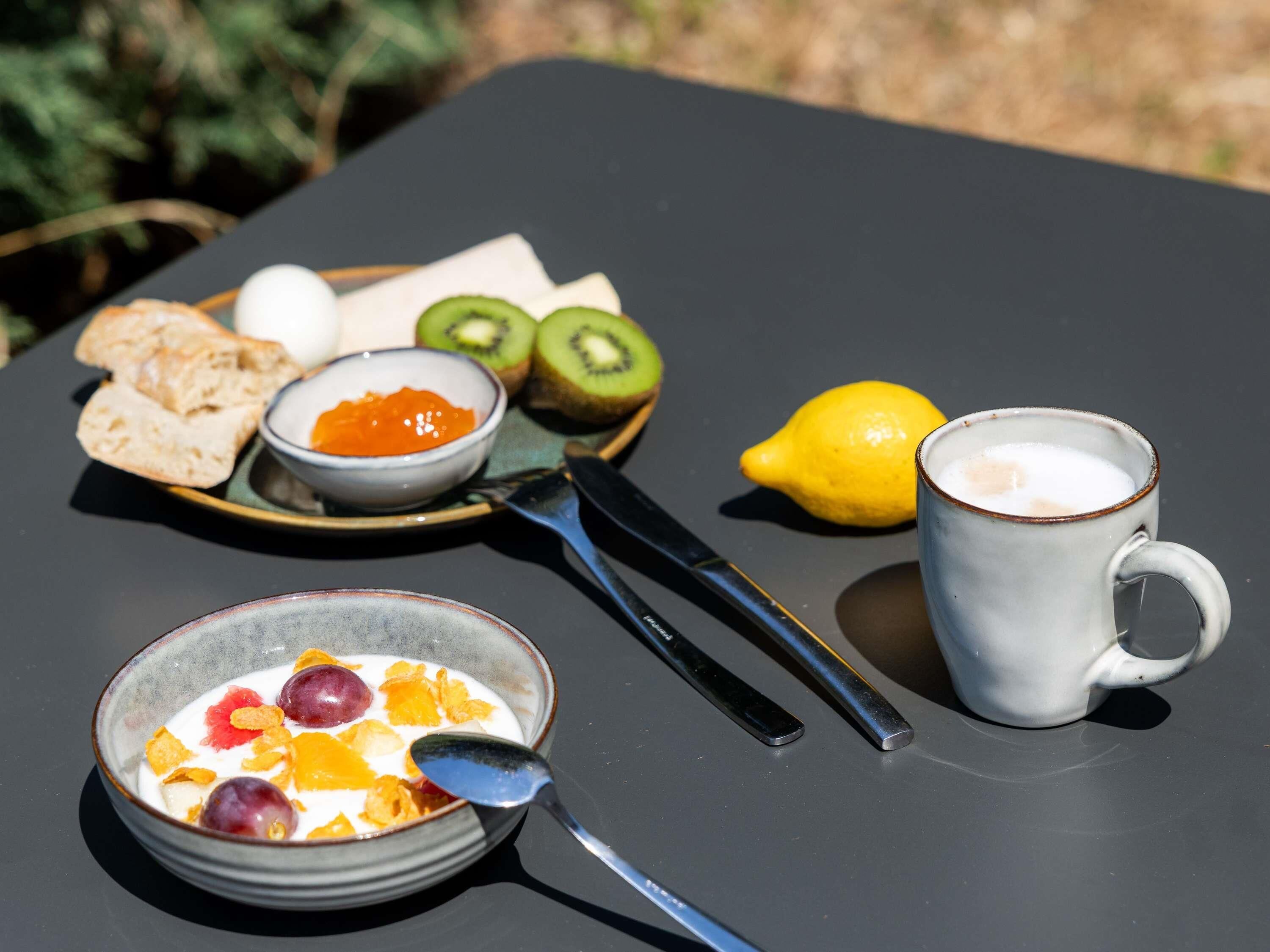 A breakfast table with a bowl of cereal, a cup of coffee, a plate with toast, kiwi, lemon, and a bowl of jam.