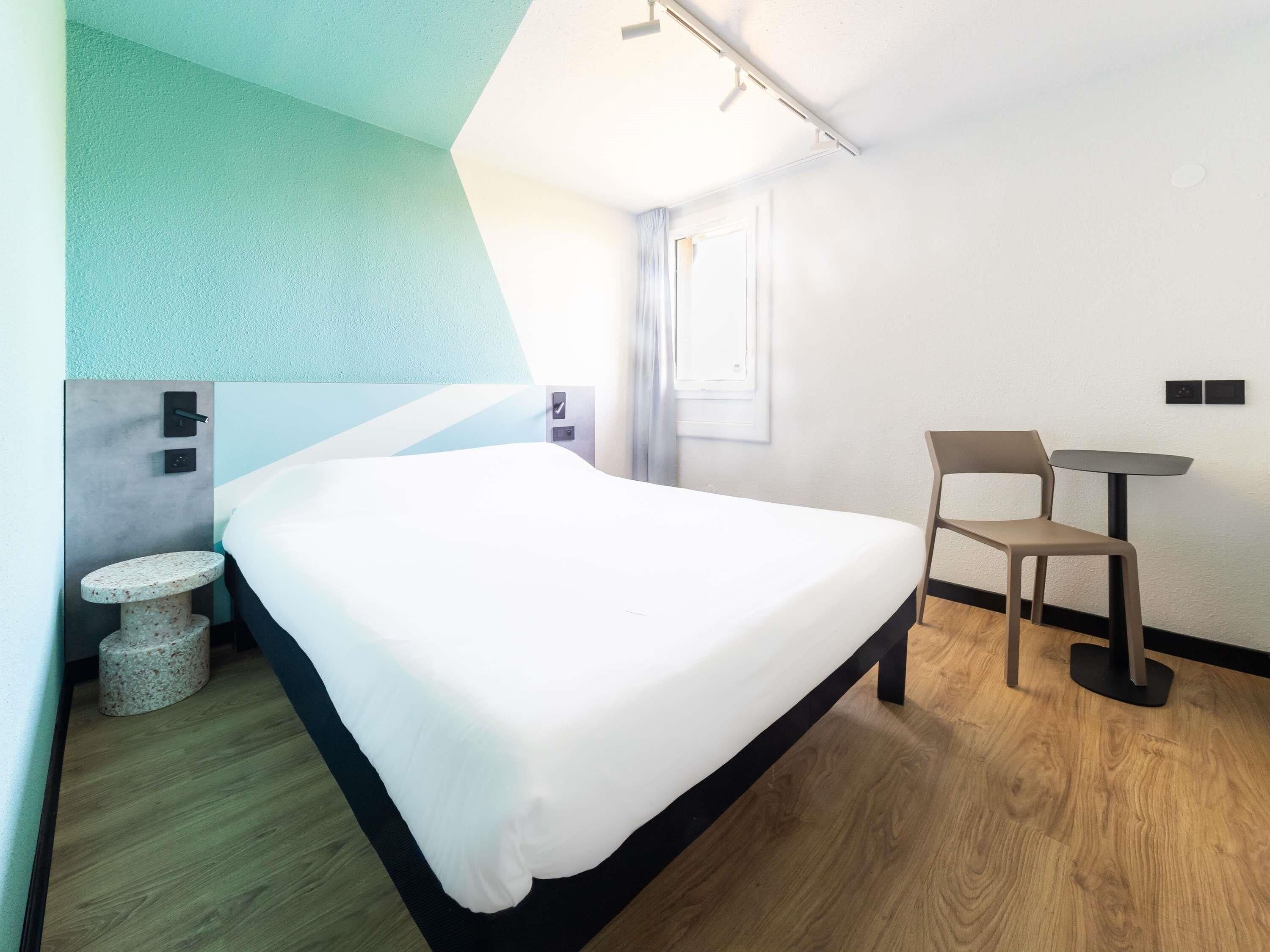 A modern hotel room with a bed, a small round table, two chairs, a window, and a door.