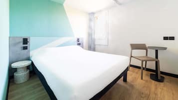 A modern hotel room with a bed, a small round table, two chairs, a window, and a door.
