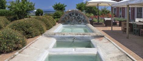 A stone water feature with a fountain, surrounded by greenery and a building with outdoor seating.