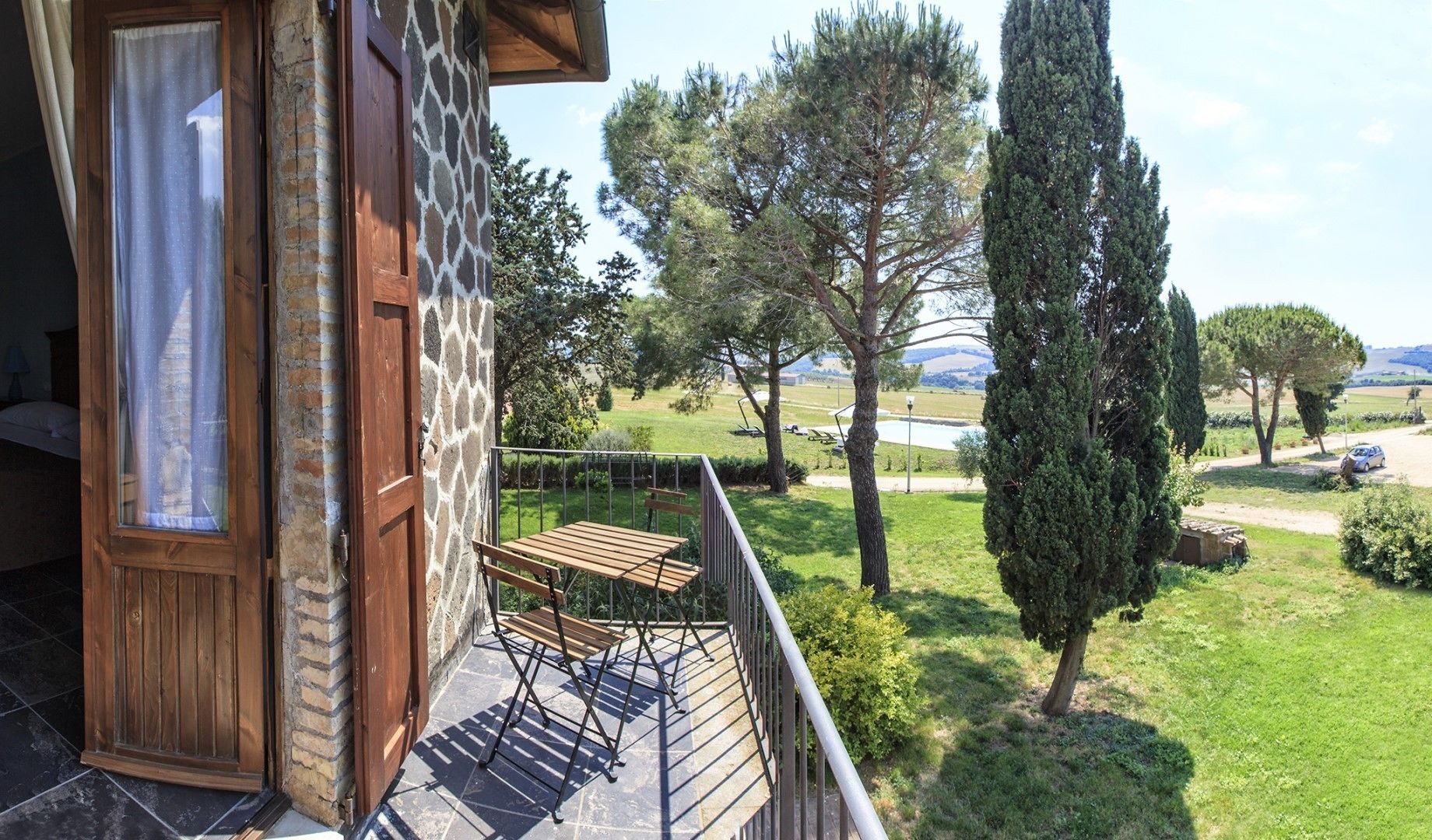 Un balcón con mesa y sillas de madera, con vistas a una zona verde con árboles y una lejana vista de colinas.