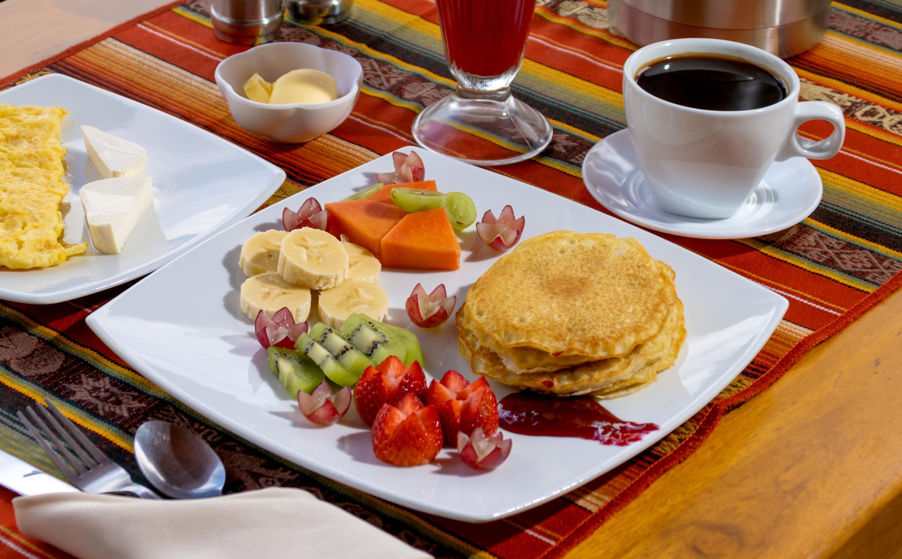 A breakfast plate with scrambled eggs, cheese, fruit, and pancakes on a colorful tablecloth.