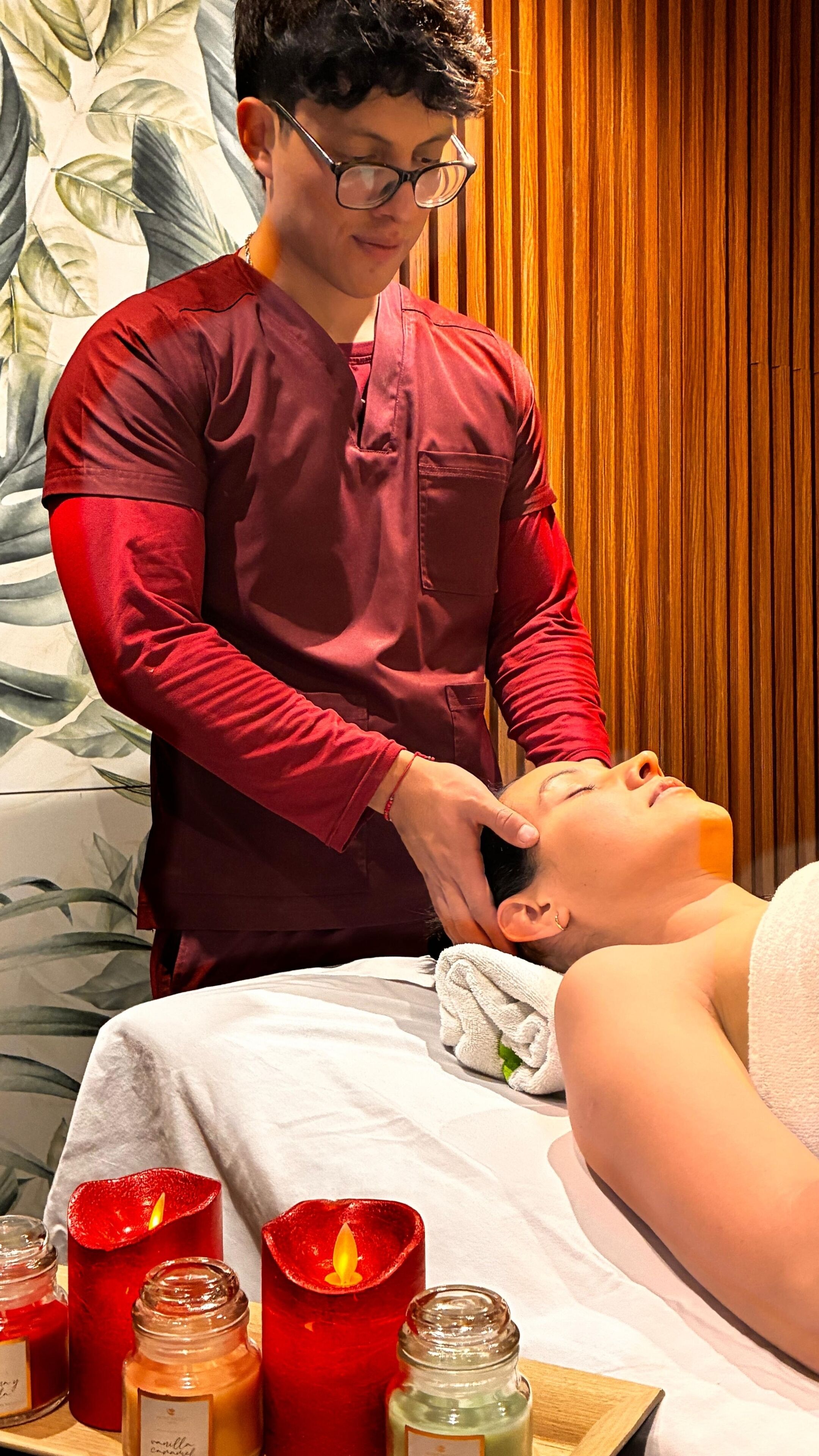 A person receiving a massage in a room with a wooden panel wall, a floral wallpaper, and several candles on a table.