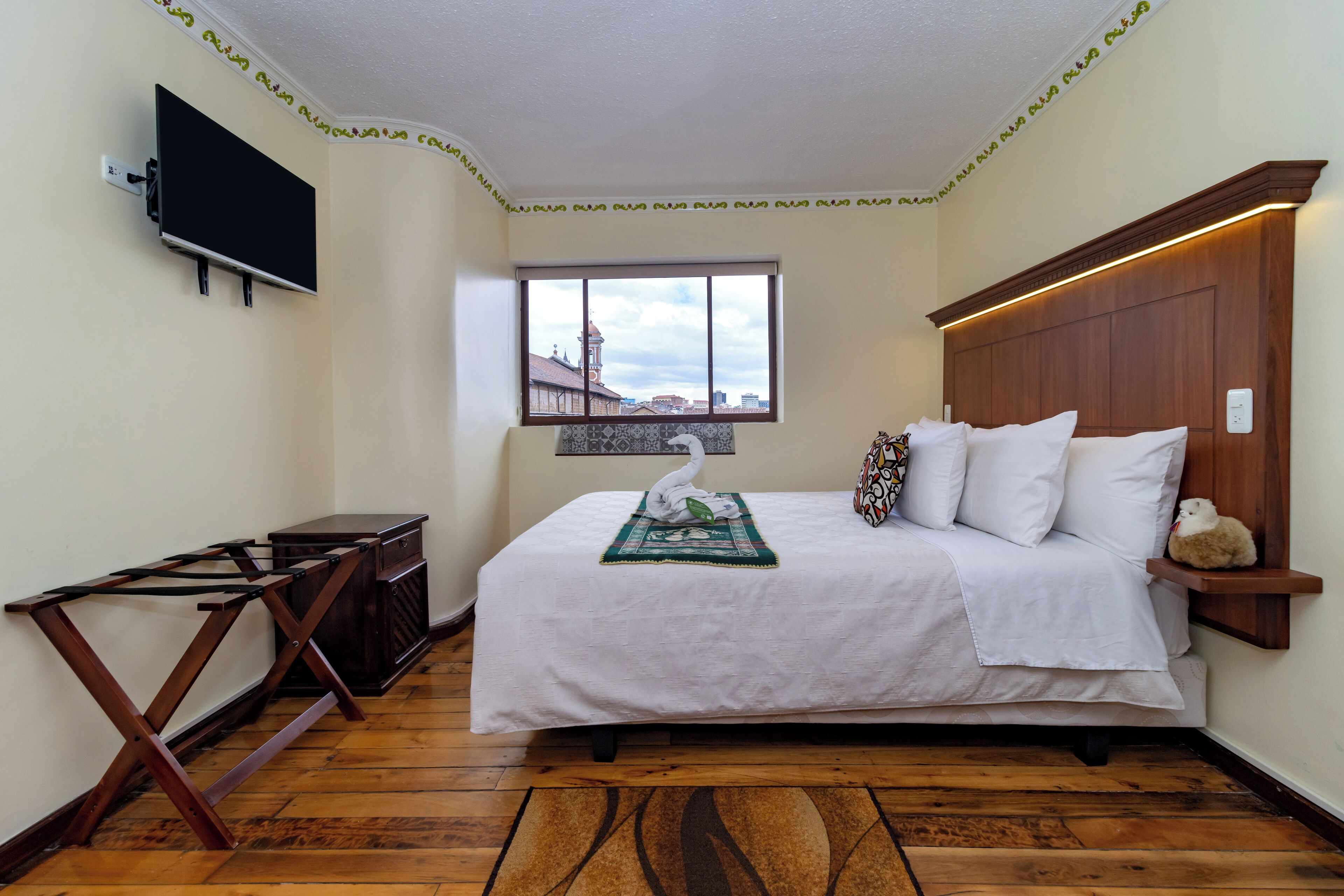 Ein Schlafzimmer mit Bett, einem an der Wand befestigten Fernseher, einem Fenster mit Blick auf die Stadt und Holzboden.