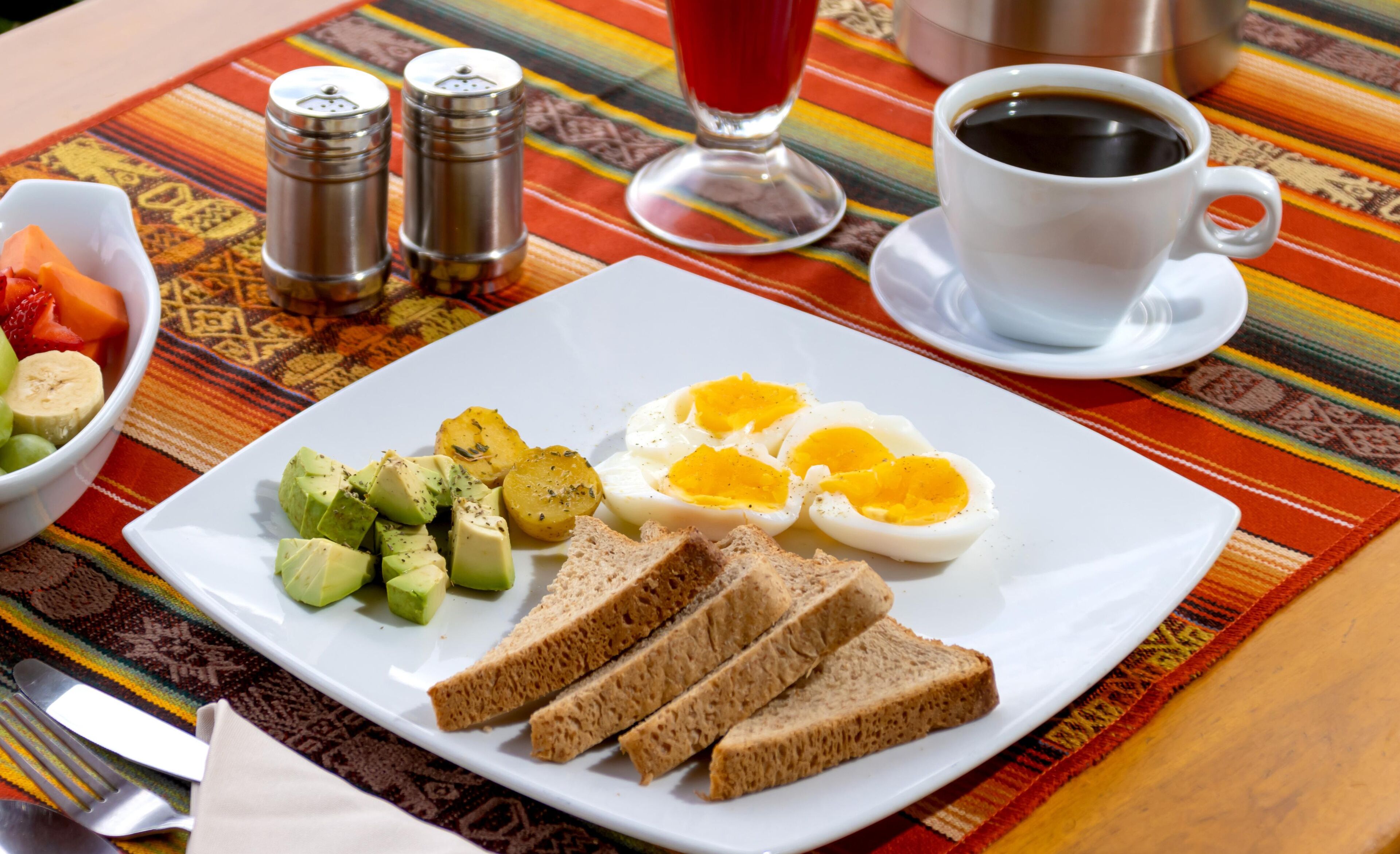 A breakfast plate with eggs, avocado, potatoes, and bread on a table with a colorful tablecloth.