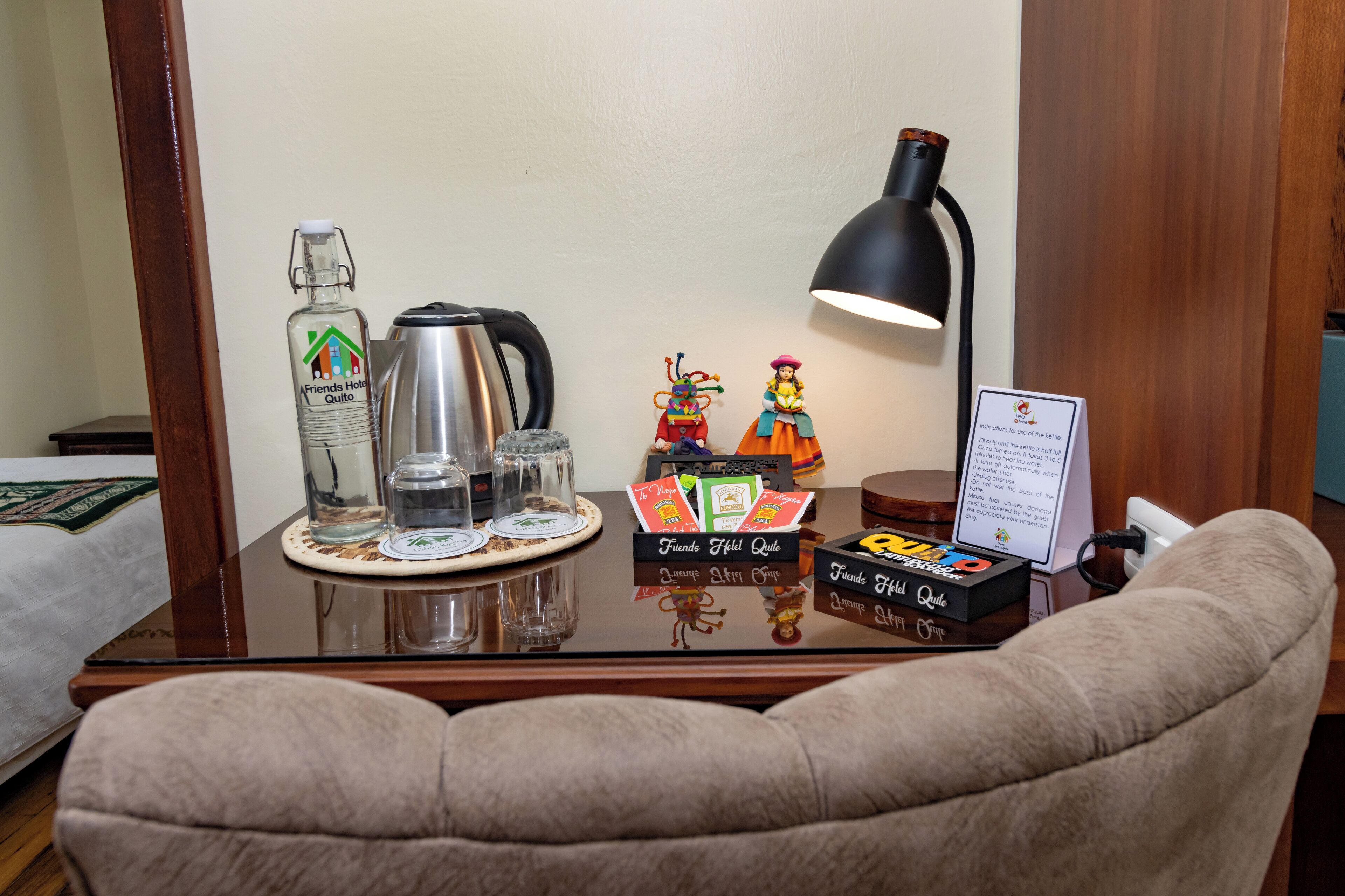Hotelværelse med et bord med glasplade, der viser en flaske spiritus, en kaffemaskine, en lampe og en lille figur.
