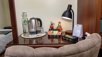 A hotel room with a glass-top table featuring a bottle of liquor, a coffee pot, a lamp, and a small figurine.